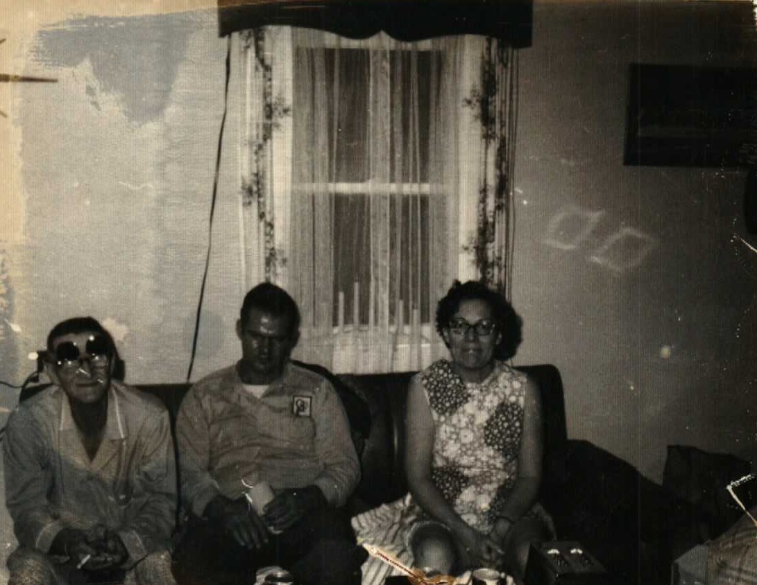 Elmer and Mary Doan sitting on a couch with an unknown man, Elmer wearing pajamas and sunglasses