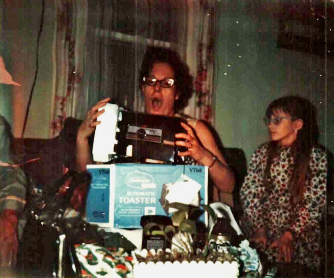 Mary Doan reacting with surprise and joy while opening a toaster at Christmas. Young Rebecca Rogers sits nearby watching